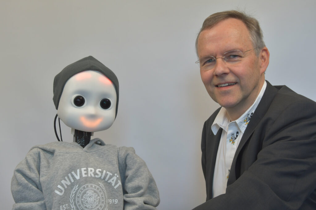 Das Foto zeigt Professor Stefan Wermter neben einem Roboter in einem Pullover mit Universitätslogo, der aussieht als würde er lächeln.